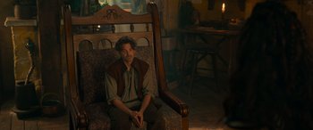 Movie still from “Dungeons & Dragons: Honor Among Thieves” (2023), directed by John Francis Daley – A man sitting on top of a couch next to a wooden chair; Medium shot, Over the shoulder angle