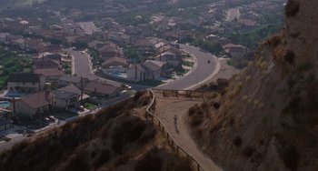 Movie still from “E.T. the Extra-Terrestrial” (1982), directed by Steven Spielberg – A person riding a bike on a dirt road near a city; Extreme Wide shot, High angle
