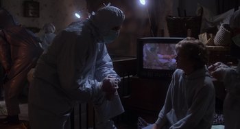 Movie still from “E.T. the Extra-Terrestrial” (1982), directed by Steven Spielberg – A man in a surgical mask is holding his hands while a woman watches television; Medium shot, Over the shoulder angle