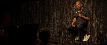 Movie still from “Chappelle's Home Team - Earthquake: Legendary” (2022), directed by Stan Lathan – A man sitting on a chair in front of an audience; Medium shot, Over the shoulder angle