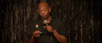 Movie still from “Chappelle's Home Team - Earthquake: Legendary” (2022), directed by Stan Lathan – A bald man holding a microphone and a cigarette; Close Up shot, Low angle