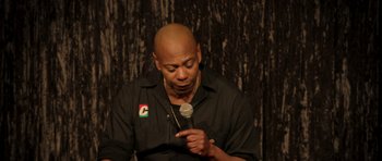 Movie still from “Chappelle's Home Team - Earthquake: Legendary” (2022), directed by Stan Lathan – A bald man holding a microphone in front of a curtain; Close Up shot, Low angle