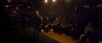 Movie still from “Chappelle's Home Team - Earthquake: Legendary” (2022), directed by Stan Lathan – A group of people sitting in a room watching a man stand up; Wide shot, Low angle