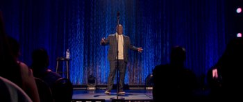 Movie still from “Chappelle's Home Team - Earthquake: Legendary” (2022), directed by Stan Lathan – A man standing on a stage holding a microphone; Wide shot, Low angle