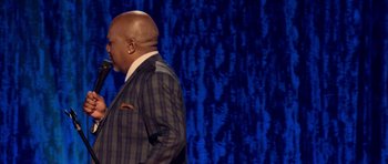Movie still from “Chappelle's Home Team - Earthquake: Legendary” (2022), directed by Stan Lathan – A man wearing a suit and tie standing in front of a blue curtain; Medium shot, Over the shoulder angle