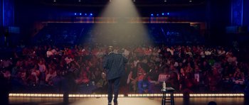 Movie still from “Chappelle's Home Team - Earthquake: Legendary” (2022), directed by Stan Lathan – A man standing on a stage in front of an audience; Extreme Wide shot, High angle