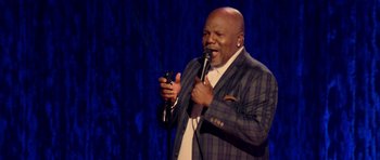 Movie still from “Chappelle's Home Team - Earthquake: Legendary” (2022), directed by Stan Lathan – A man holding a cell phone while standing on a stage; Medium shot, Low angle