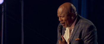 Movie still from “Chappelle's Home Team - Earthquake: Legendary” (2022), directed by Stan Lathan – A man with a shaved head is holding a microphone; Close Up shot, Low angle