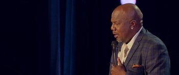 Movie still from “Chappelle's Home Team - Earthquake: Legendary” (2022), directed by Stan Lathan – A man holding onto a microphone while standing in front of a curtain; Close Up shot, Over the shoulder angle