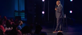 Movie still from “Chappelle's Home Team - Earthquake: Legendary” (2022), directed by Stan Lathan – A man standing on a stage holding a microphone; Wide shot, Low angle