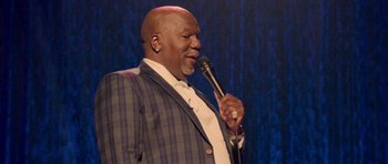 Movie still from “Chappelle's Home Team - Earthquake: Legendary” (2022), directed by Stan Lathan – A man in a suit and tie holding a microphone; Medium shot, Over the shoulder angle