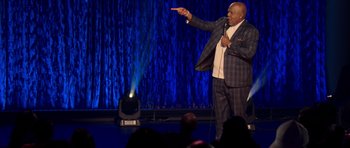 Movie still from “Chappelle's Home Team - Earthquake: Legendary” (2022), directed by Stan Lathan – A man standing on a stage holding a microphone; Medium shot, Over the shoulder angle
