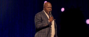 Movie still from “Chappelle's Home Team - Earthquake: Legendary” (2022), directed by Stan Lathan – A man in a suit and tie holding a microphone; Medium shot, Low angle