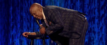 Movie still from “Chappelle's Home Team - Earthquake: Legendary” (2022), directed by Stan Lathan – A man in a suit and tie holding a microphone; Medium shot, High angle