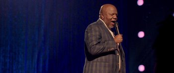 Movie still from “Chappelle's Home Team - Earthquake: Legendary” (2022), directed by Stan Lathan – A man in a suit and tie holding a microphone and singing; Medium shot, Low angle