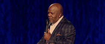 Movie still from “Chappelle's Home Team - Earthquake: Legendary” (2022), directed by Stan Lathan – A man holding a microphone while wearing a suit; Close Up shot, High angle