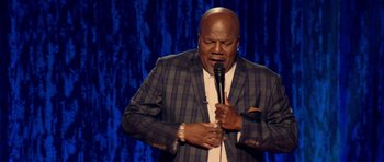 Movie still from “Chappelle's Home Team - Earthquake: Legendary” (2022), directed by Stan Lathan – A man holding a microphone while standing on a stage; Medium shot, Low angle