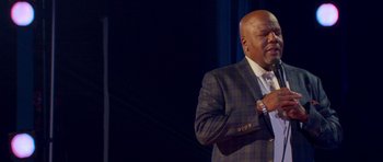 Movie still from “Chappelle's Home Team - Earthquake: Legendary” (2022), directed by Stan Lathan – A man standing on stage with a microphone in front of a crowd; Medium shot, Low angle