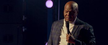 Movie still from “Chappelle's Home Team - Earthquake: Legendary” (2022), directed by Stan Lathan – An older man holding a microphone while standing up; Medium shot, Low angle