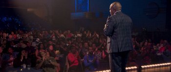Movie still from “Chappelle's Home Team - Earthquake: Legendary” (2022), directed by Stan Lathan – A man standing in front of a crowd of people in a room; Wide shot, Over the shoulder angle