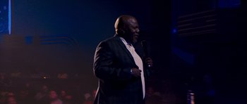 Movie still from “Chappelle's Home Team - Earthquake: Legendary” (2022), directed by Stan Lathan – A man holding a microphone while wearing a suit and tie; Medium shot, Low angle
