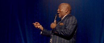 Movie still from “Chappelle's Home Team - Earthquake: Legendary” (2022), directed by Stan Lathan – A man holding a microphone while standing on a stage; Medium shot, Low angle