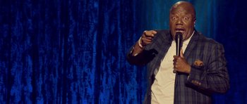Movie still from “Chappelle's Home Team - Earthquake: Legendary” (2022), directed by Stan Lathan – A man standing on a stage holding a microphone; Medium shot, Over the shoulder angle