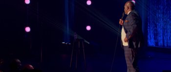 Movie still from “Chappelle's Home Team - Earthquake: Legendary” (2022), directed by Stan Lathan – A man standing in front of a microphone on a stage; Wide shot, High angle