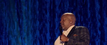 Movie still from “Chappelle's Home Team - Earthquake: Legendary” (2022), directed by Stan Lathan – A man holding a microphone while standing in front of a blue curtain; Medium shot, Over the shoulder angle