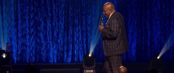 Movie still from “Chappelle's Home Team - Earthquake: Legendary” (2022), directed by Stan Lathan – A man holding a microphone while standing on a stage; Medium shot, Over the shoulder angle