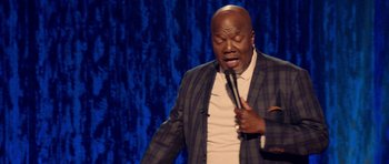 Movie still from “Chappelle's Home Team - Earthquake: Legendary” (2022), directed by Stan Lathan – A man holding a microphone while standing on a stage; Medium shot, High angle