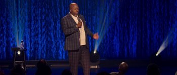 Movie still from “Chappelle's Home Team - Earthquake: Legendary” (2022), directed by Stan Lathan – A man standing on a stage holding a microphone; Medium shot, Over the shoulder angle