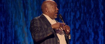 Movie still from “Chappelle's Home Team - Earthquake: Legendary” (2022), directed by Stan Lathan – A man in a suit and tie holding a microphone; Medium shot, Low angle