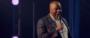 Movie still from “Chappelle's Home Team - Earthquake: Legendary” (2022), directed by Stan Lathan – A man in a suit and tie holding a microphone; Medium shot, Low angle