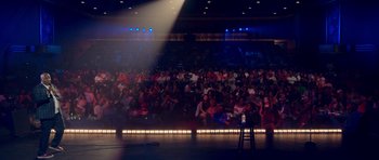 Movie still from “Chappelle's Home Team - Earthquake: Legendary” (2022), directed by Stan Lathan – A crowd of people sitting on a stage in front of a microphone; Extreme Wide shot, High angle