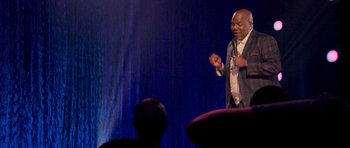 Movie still from “Chappelle's Home Team - Earthquake: Legendary” (2022), directed by Stan Lathan – A man standing on a stage holding a microphone; Medium shot, Over the shoulder angle