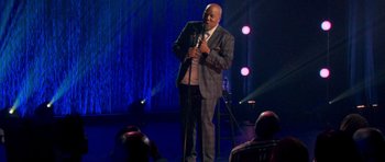 Movie still from “Chappelle's Home Team - Earthquake: Legendary” (2022), directed by Stan Lathan – A man standing on a stage holding a microphone; Wide shot, Over the shoulder angle