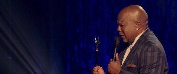 Movie still from “Chappelle's Home Team - Earthquake: Legendary” (2022), directed by Stan Lathan – A man holding a microphone on a stage; Close Up shot, High angle