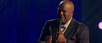 Movie still from “Chappelle's Home Team - Earthquake: Legendary” (2022), directed by Stan Lathan – A man holding a microphone while wearing a suit; Close Up shot, High angle