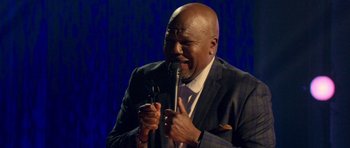 Movie still from “Chappelle's Home Team - Earthquake: Legendary” (2022), directed by Stan Lathan – A man in a suit and tie holding a microphone; Medium shot, High angle