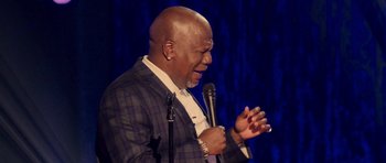 Movie still from “Chappelle's Home Team - Earthquake: Legendary” (2022), directed by Stan Lathan – A bald man in a blue jacket is holding a microphone; Medium shot, Over the shoulder angle