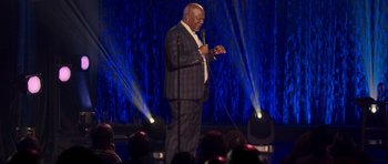 Movie still from “Chappelle's Home Team - Earthquake: Legendary” (2022), directed by Stan Lathan – A man standing on a stage holding a microphone; Medium shot, Low angle