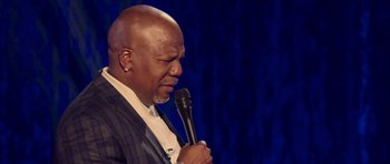 Movie still from “Chappelle's Home Team - Earthquake: Legendary” (2022), directed by Stan Lathan – A man holding a microphone while wearing a suit; Close Up shot, Over the shoulder angle