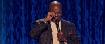 Movie still from “Chappelle's Home Team - Earthquake: Legendary” (2022), directed by Stan Lathan – A man holding onto a microphone while holding his hand up to his face; Close Up shot, High angle