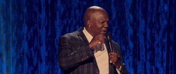 Movie still from “Chappelle's Home Team - Earthquake: Legendary” (2022), directed by Stan Lathan – A man in a suit and bow tie holding a microphone; Medium shot, Over the shoulder angle