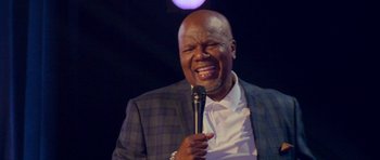 Movie still from “Chappelle's Home Team - Earthquake: Legendary” (2022), directed by Stan Lathan – A man with a microphone in his hand; Close Up shot, Low angle