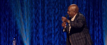 Movie still from “Chappelle's Home Team - Earthquake: Legendary” (2022), directed by Stan Lathan – A man holding a microphone while standing on a stage; Medium shot, High angle