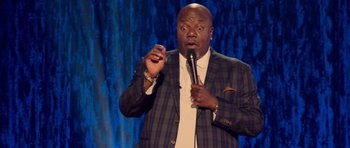 Movie still from “Chappelle's Home Team - Earthquake: Legendary” (2022), directed by Stan Lathan – A man holding a microphone in front of a microphone stand; Medium shot, High angle