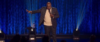Movie still from “Chappelle's Home Team - Earthquake: Legendary” (2022), directed by Stan Lathan – A man standing on a stage holding a microphone; Medium shot, Low angle