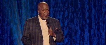 Movie still from “Chappelle's Home Team - Earthquake: Legendary” (2022), directed by Stan Lathan – A man holding a microphone while standing on a stage; Close Up shot, Low angle